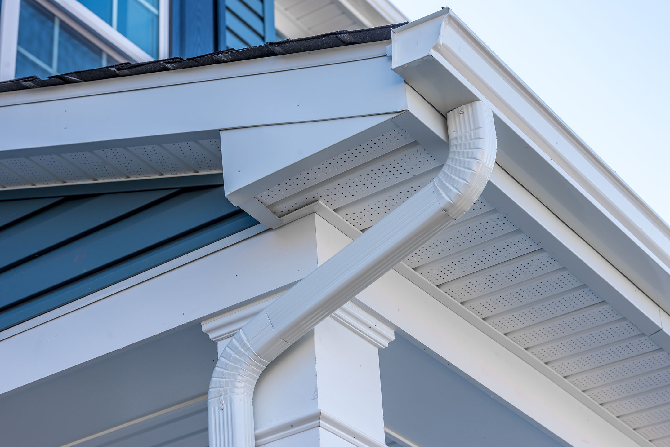 Close-up of a white gutter system on a modern house. The gutter is attached to a roof with blue siding. The soffit and fascia are also white, complementing the overall aesthetic of the home. The sky is clear in the background.