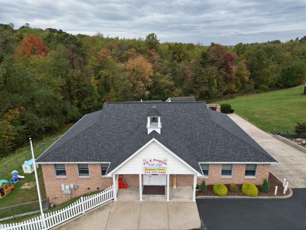 A single-story brick building with a dark gray roof, surrounded by a large lawn and leafy autumn trees. A playground set is visible to the left. There's a long driveway, and a sign displayed on the building's front. The sky is overcast.