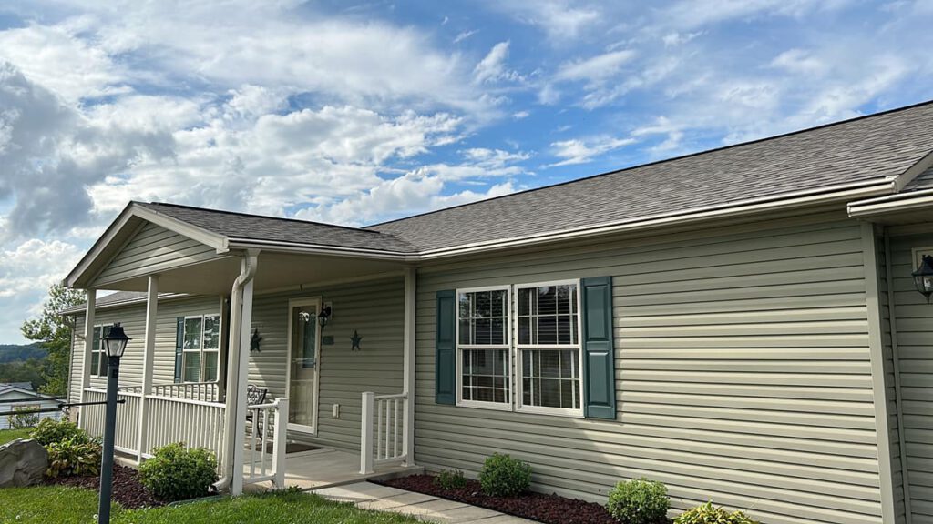 A single-story house with beige siding and green shutters. It features a spacious porch with white railings, surrounded by a manicured lawn and shrubs. The sky above is partly cloudy with patches of blue.