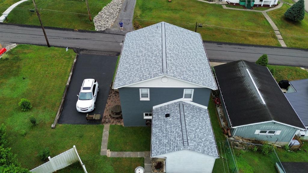Aerial view of a suburban area featuring a two-story gray house with a gray roof. A white car is parked in the driveway. The surrounding area includes green lawns and intersecting roads. Another house with a dark roof is adjacent.
