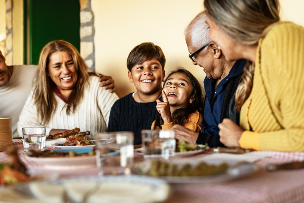 A family gathered around a table, smiling and laughing. A young girl is looking up joyfully with a boy beside her. An older man in glasses leans toward the group. The table is set with food and drinks.