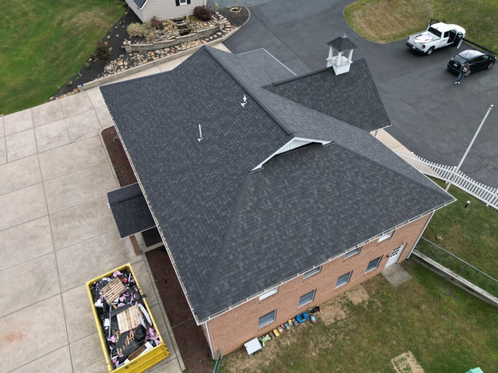 Aerial view of a building with a dark gray roof, surrounded by a driveway and grassy area. Two vehicles are parked nearby. A yellow dumpster, partially filled with materials, is visible at the bottom of the image.